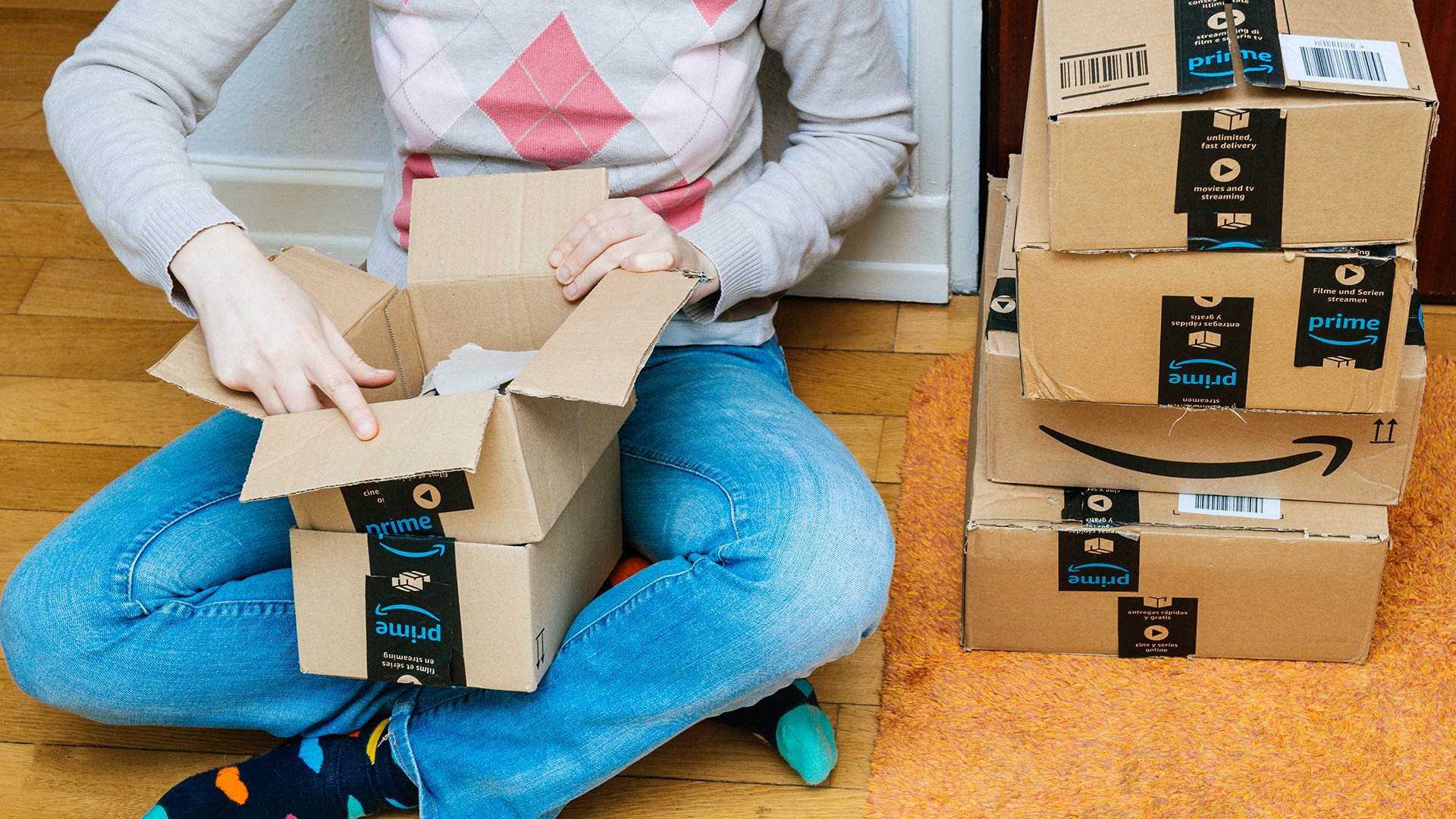 A person sitting on the floor next to a stack of Amazon boxes and opening one.