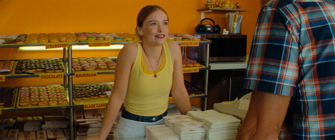 A girl in a yellow top smiles while leaning over donut shop counter in 'Red Rocket.'