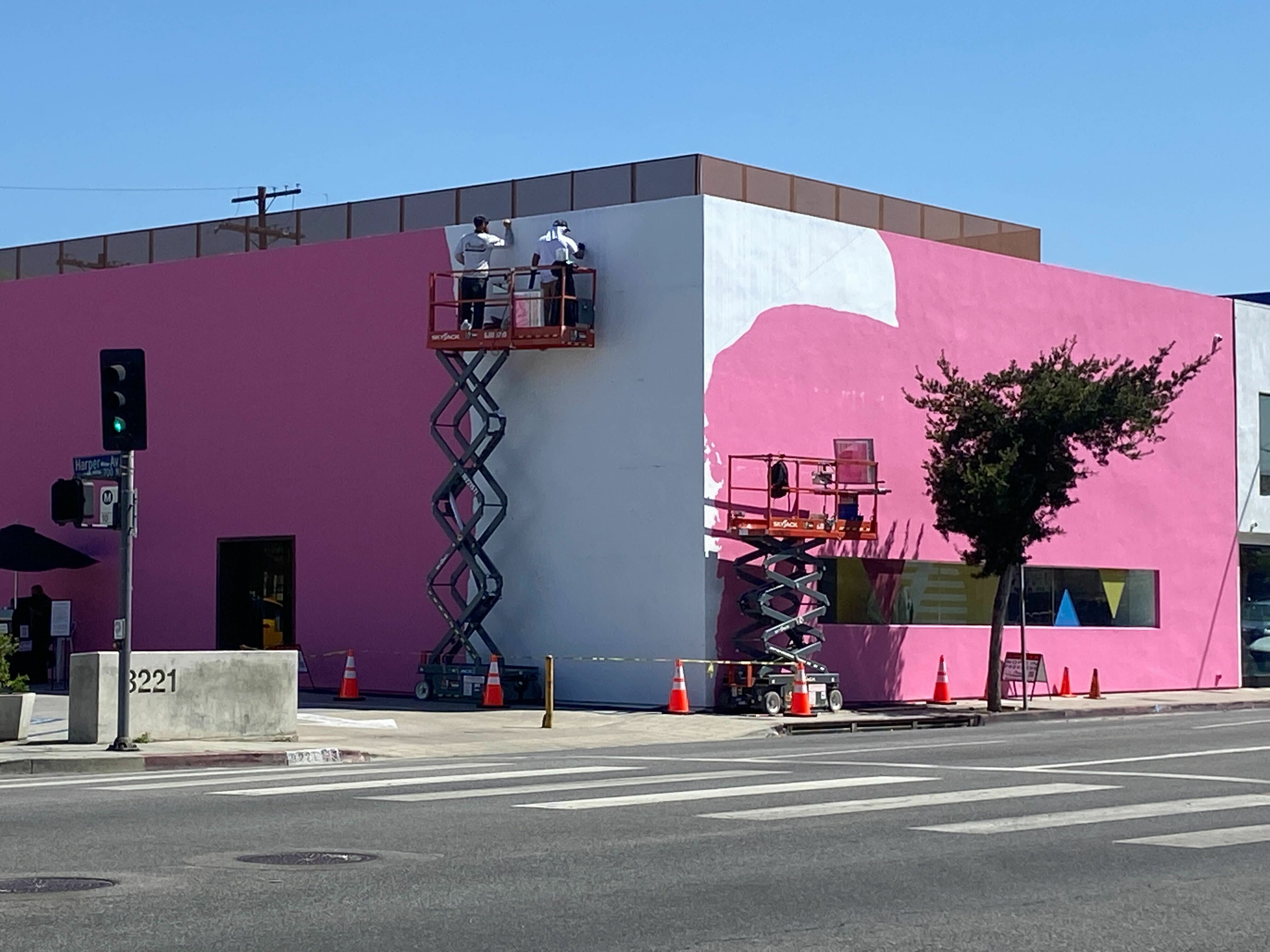 A pink building with painters painting the corner white.