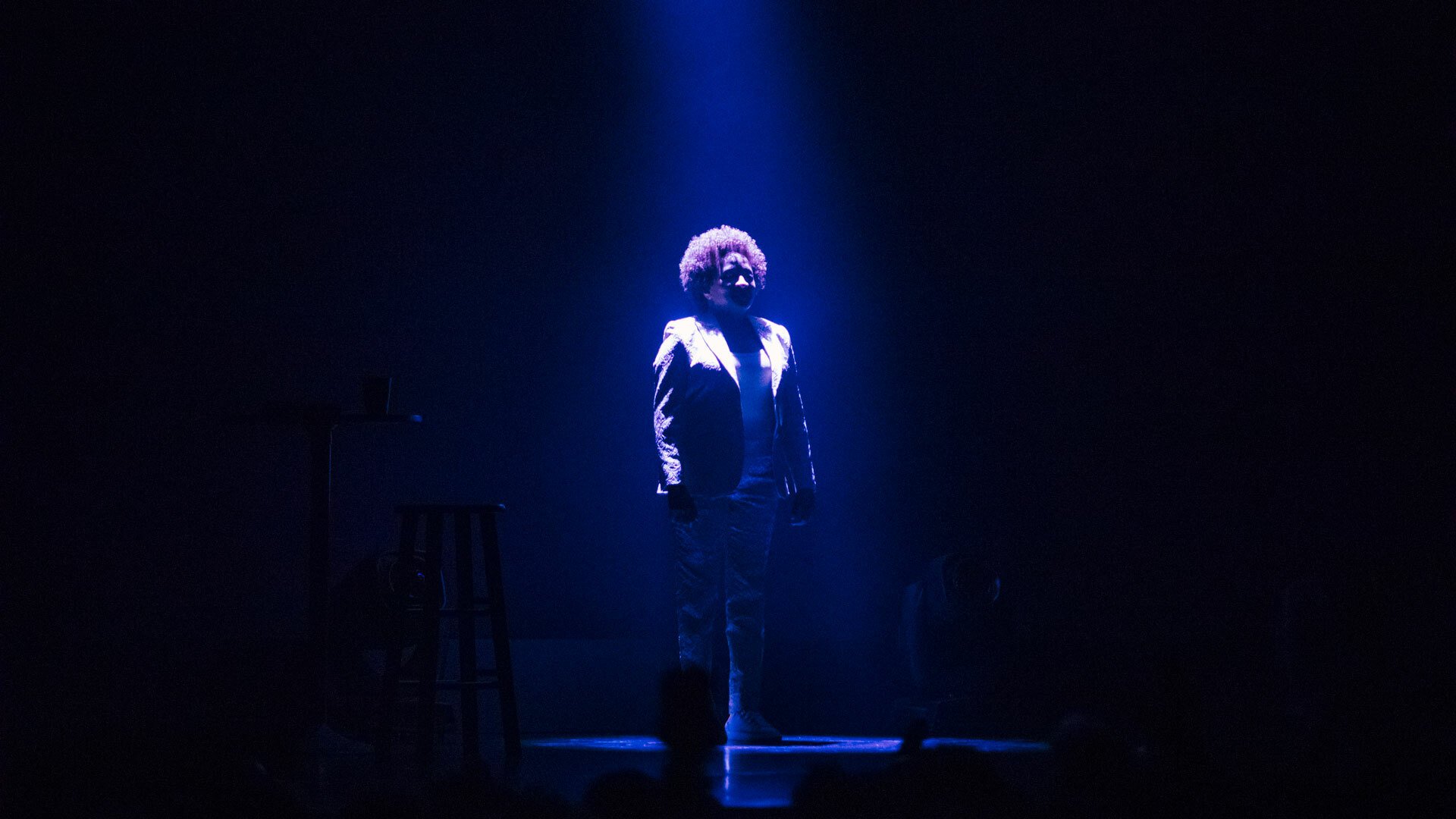 Wanda Sykes stands on stage, dressed in a white suit and bathed in the light of a blue spotlight.