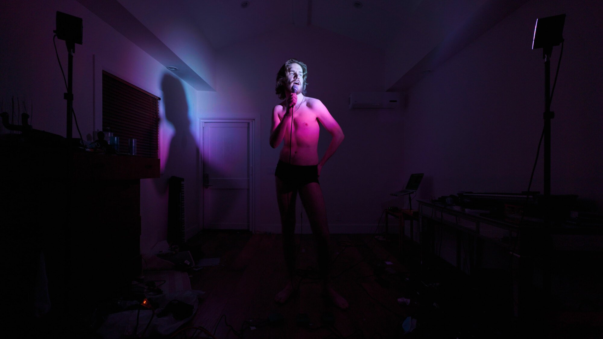 Bo Burnham stands in an apartment bathed in purplish light. He's holding a microphone and not wearing any clothes.