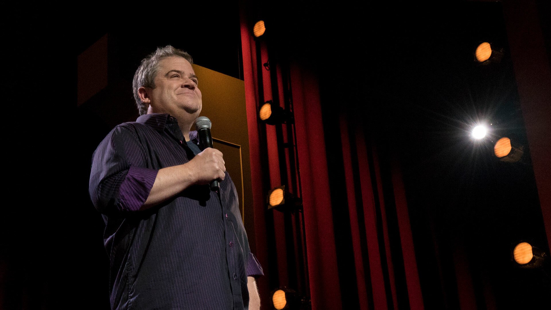 Patton Oswalt stands on a stage holding a microphone.