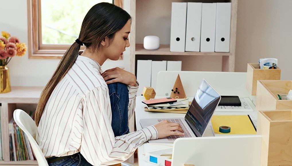 Woman working on laptop at desk with Google Nest mesh WiFi router on bookshelf behind her