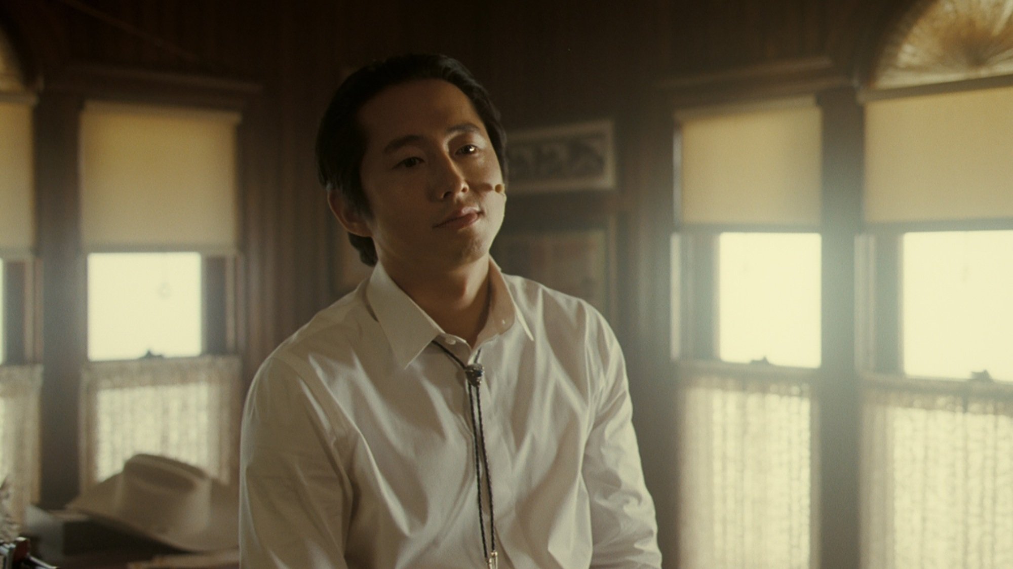 A man in a white button up and a bolo tie sits on an office desk, staring off into space. A white cowboy hat is on the desk behind him.