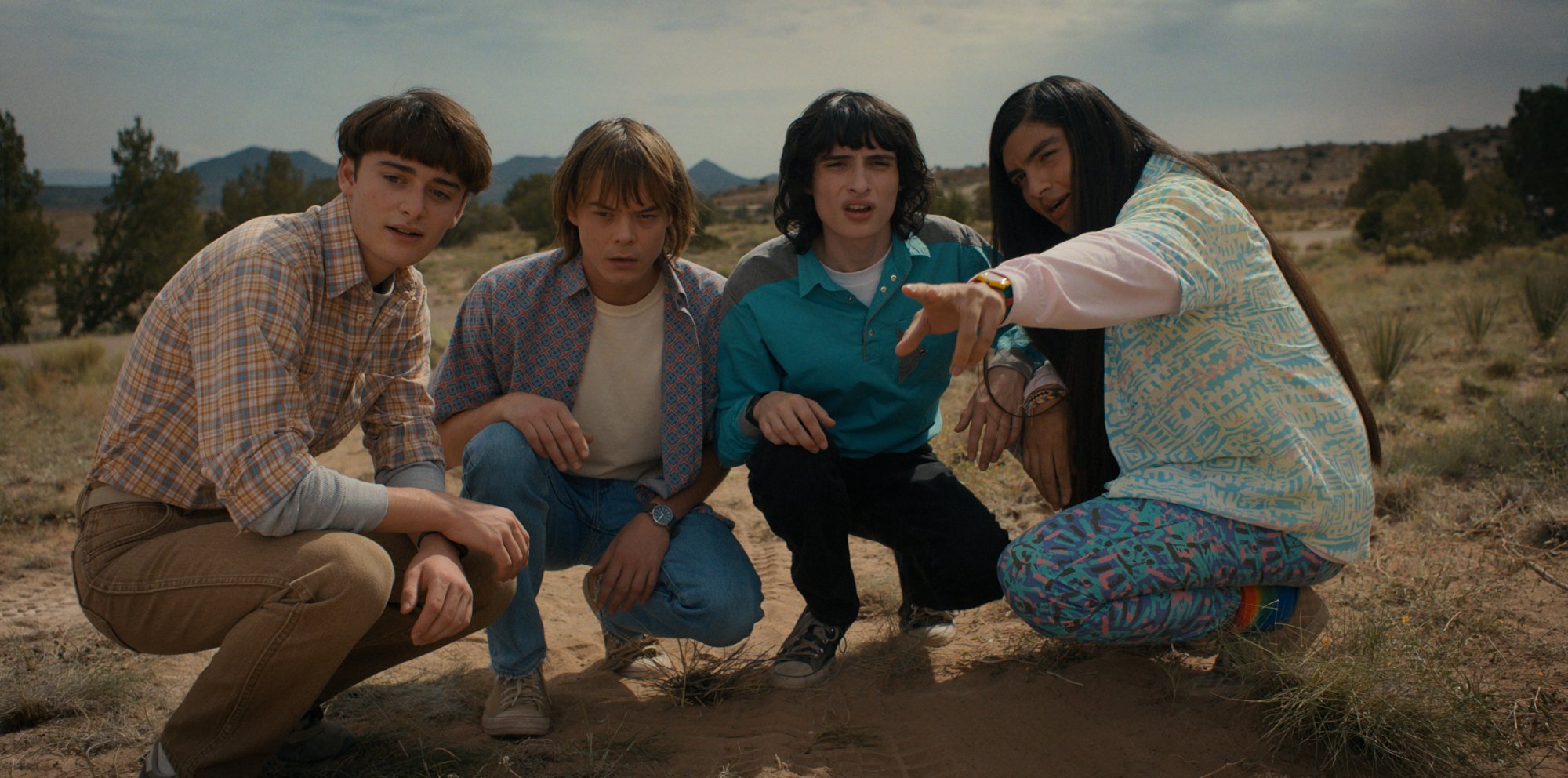 Four young men squat in the desert, one pointing towards something in the distance.