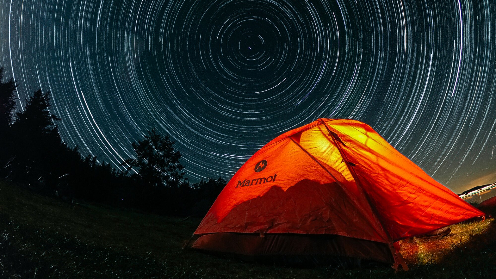 A photograph of star trails in the night sky over an orange tent.