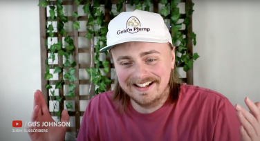 A screenshot of a Gus Johnson video. Johnson appears to be sitting, as you can only see him from the chest up. He is wearing a while baseball hat and a red shirt in front of a trellis of green plants behind him.